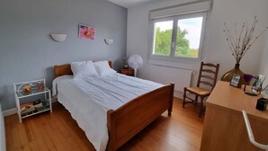 A bedroom with a wooden bed, a nightstand with a fan, a chair, a table with a vase, and a window with a view of greenery. - Country house with wonderful view over the Lot valley (Galapian)