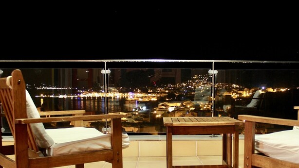 Ein Balkon mit zwei Holzstühlen und einem kleinen Tisch, mit Blick auf die nächtliche Skyline.