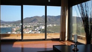 Ein geräumiges Zimmer mit großen Fenstern und Blick auf eine Küstenstadt und Berge.