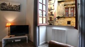 Un salon avec un téléviseur posé sur un meuble blanc, un tableau accroché au mur et une fenêtre donnant sur de vieux bâtiments.