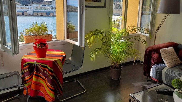 A room with a view of a marina, a table with a colorful tablecloth, a potted plant, a sofa with a cushion, and a lamp. - Stunning fully renovated apartment with air-conditioning  (Pyrénées-Orientales)