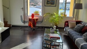 A living room with a view of a body of water and a bridge, a sofa, a coffee table with books, and a small table with a plant. - Stunning fully renovated apartment with air-conditioning  (Pyrénées-Orientales)