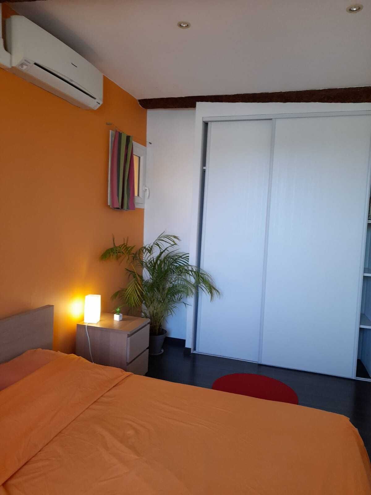 A bedroom with an orange bed, a bedside table with a lamp, a potted plant, and a sliding door to a balcony.
