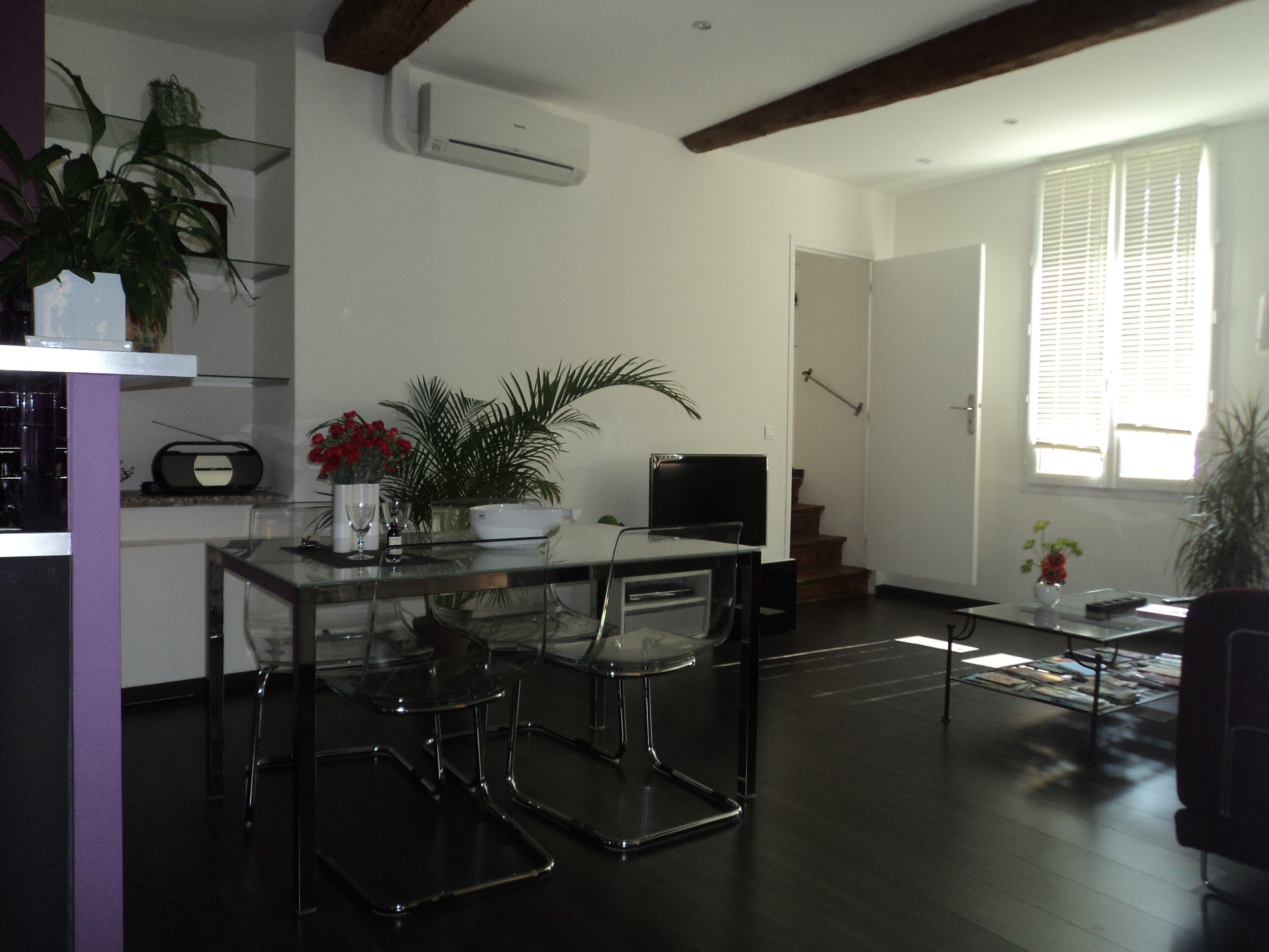 A modern living room with a glass dining table, clear chairs, a flat-screen TV, and a staircase leading to another level.