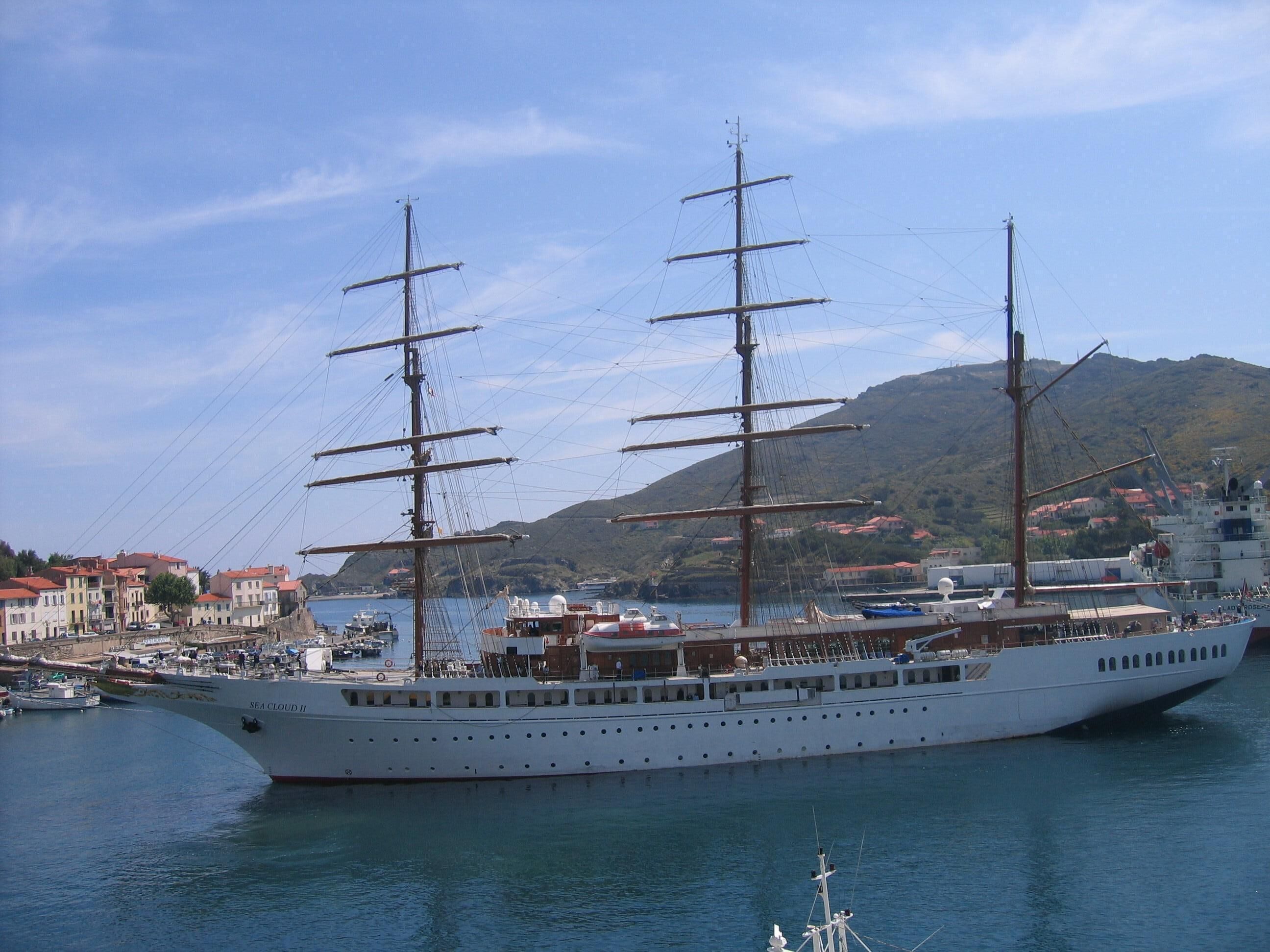 A large white cruise ship docked at a port with a mountainous backdrop and a town with red-roofed buildings.