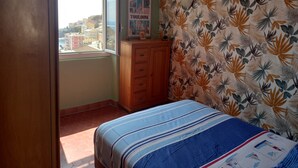 Un dormitori amb llit, un taulell de fusta, una finestra amb vista a edificis i una paret amb dibuixos de fulles.