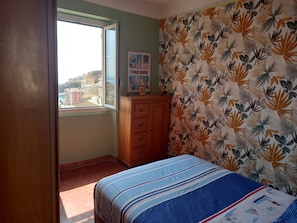 Ein Schlafzimmer mit einem Bett, einer Holzkommode, einem Fenster mit Blick auf Gebäude und einer Wand mit einem Blattmuster.