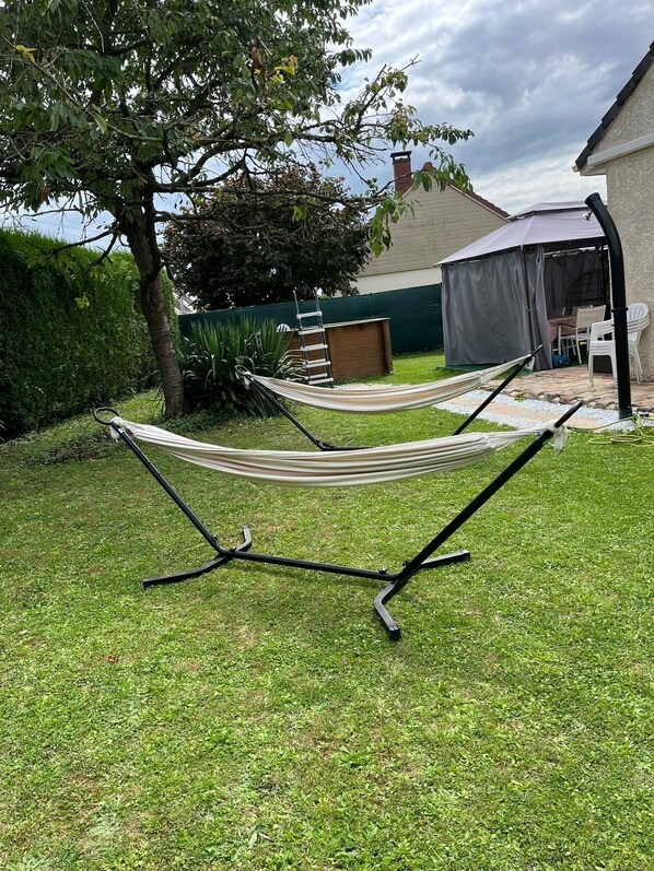 Un hamac installé dans un jardin, avec une pelouse verdoyante, un arbre et une maison en arrière-plan.