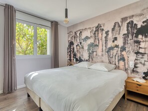 Une chambre à coucher avec un lit, une fenêtre avec des rideaux, un mur orné d’un tableau et une table de chevet avec un livre.