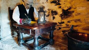 Eine Person sitzt an einem Holztisch, schreibt bei Schreibtischlampe und hat einen großen Topf in der Nähe.