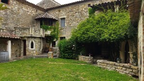 Edificio de piedra con un patio, que presenta una zona verde con flores amarillas, muros de piedra y un techo de tejas.
