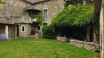 A stone building with a courtyard, featuring a grassy area with yellow flowers, stone walls, and a tiled roof.