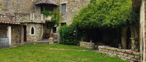 A stone building with a courtyard, featuring a grassy area with yellow flowers, stone walls, and a tiled roof.