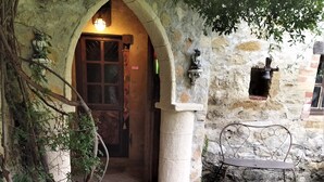Un arco de piedra al aire libre que da paso a una puerta de madera, un banco de piedra y un farol sobre una superficie de piedra.