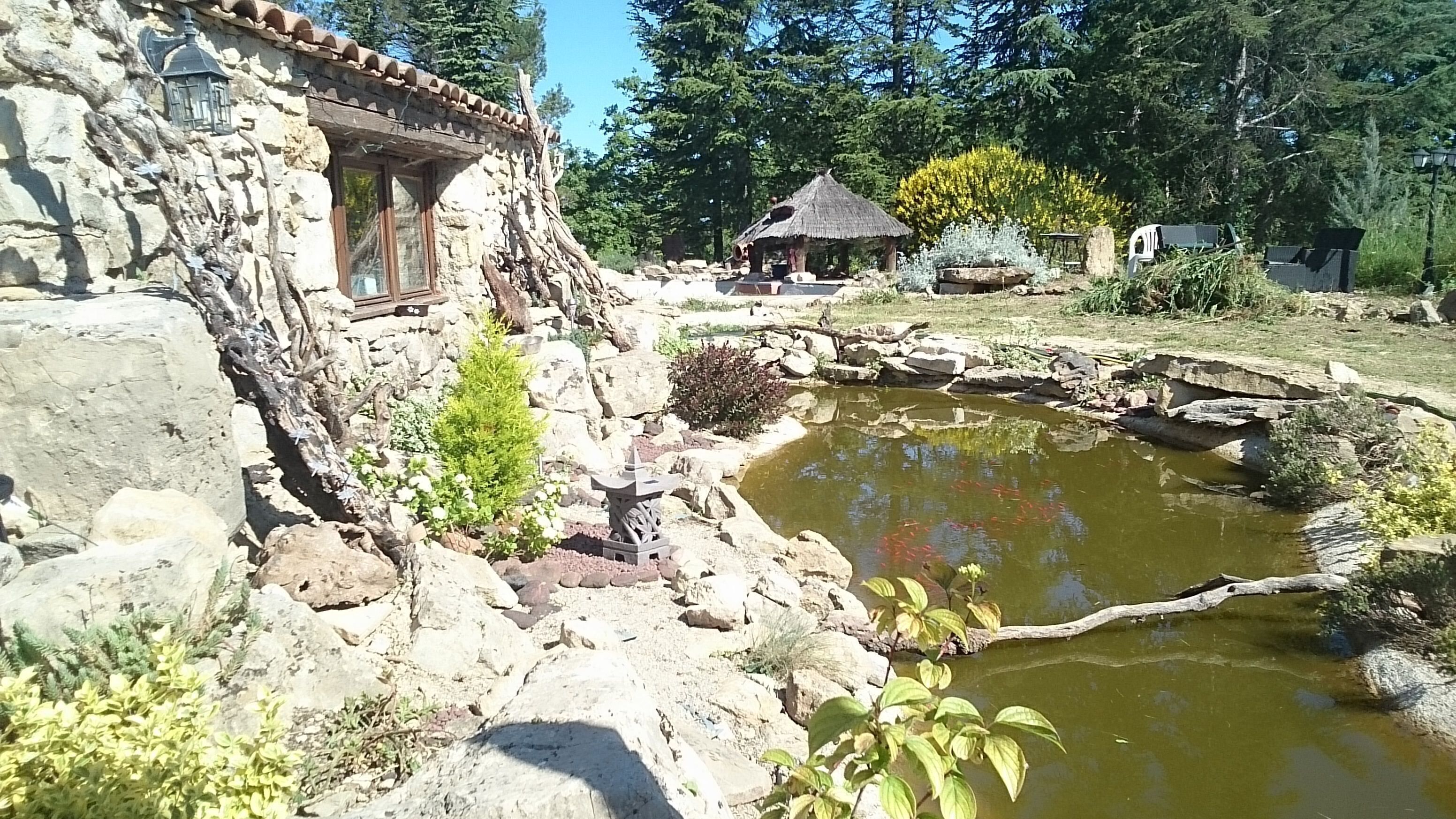 A rustic stone building with a thatched-roof structure nearby, a small pond with koi fish, and surrounding greenery.