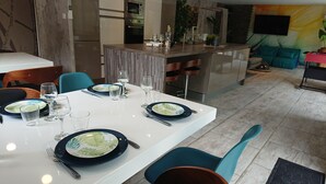 A modern dining area with a white table set for six, blue chairs, and a kitchen in the background.