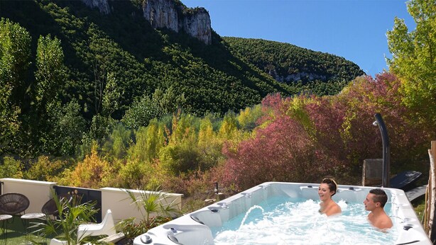 A hot tub with two people in it, surrounded by greenery and a mountain in the background.
