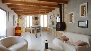 A cozy living room with a white sofa, a white armchair, and a dining area with a table and chairs. There is a fireplace, stone walls, and large windows. - IDEAL COZY HOUSE FOR 1 COUPLE AND 2 CHILDREN  (Sainte-Marie-de-Ré)