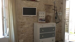 A room with a checkered floor, a grey sofa with a red pillow, a white dresser with drawers, a mounted television, and a wall-mounted shelf with a red object. - Charming typical house  (Meschers-sur-Gironde)