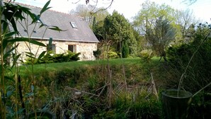 Uma casa de pedra com telhado inclinado, rodeada por um jardim exuberante e árvores densas.