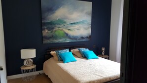 Een slaapkamer met een groot schilderij met een oceaanmotief, een bed met blauwe en beige beddengoed, een bedlamp en een klein rond tafeltje met een decoratief object.