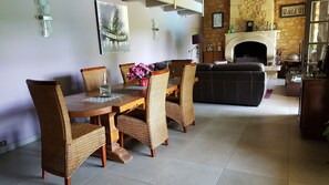 A dining area with a wooden table and wicker chairs, a sofa, and a fireplace in the background.