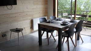 Comedor con mesa y sillas de madera, televisor montado en la pared y vistas a árboles a través de amplios ventanales.