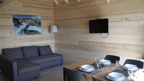 Interior de una cabaña de madera con sofá, mesa de comedor, sillas y un televisor montado.