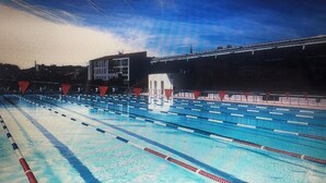 Ein Schwimmbecken mit mehreren Bahnen, im Hintergrund ein Gebäude und ein klarer Himmel.