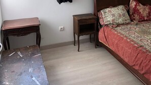 Ein Schlafzimmer mit Bett, Nachttisch, kleinem Tisch, Fenster mit Vorhang und Blick auf Bäume.