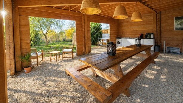 Une cabane en bois rustique, avec une grande table et des bancs en bois, un luminaire suspendu en fibres tressées et une vue sur le jardin environnant.