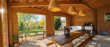 A rustic wooden cabin with a large wooden table and benches, a hanging woven light fixture, and a view of the surrounding garden.