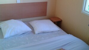 A single bed with two pillows, a headboard, a small wooden nightstand, and a window with blinds.