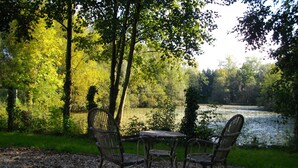 A table and four wicker chairs set on a gravel surface, surrounded by trees and overlooking a calm river.