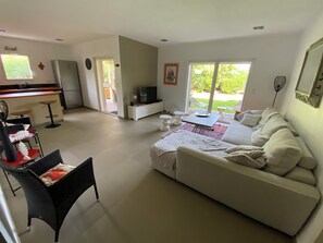 A modern living room with a sofa, a dining area with chairs, a kitchen area with a refrigerator, and a large window providing a view of the outdoors. - New luxury apartment in an apartment building, 70 m from Pinarello beach (Zonza)