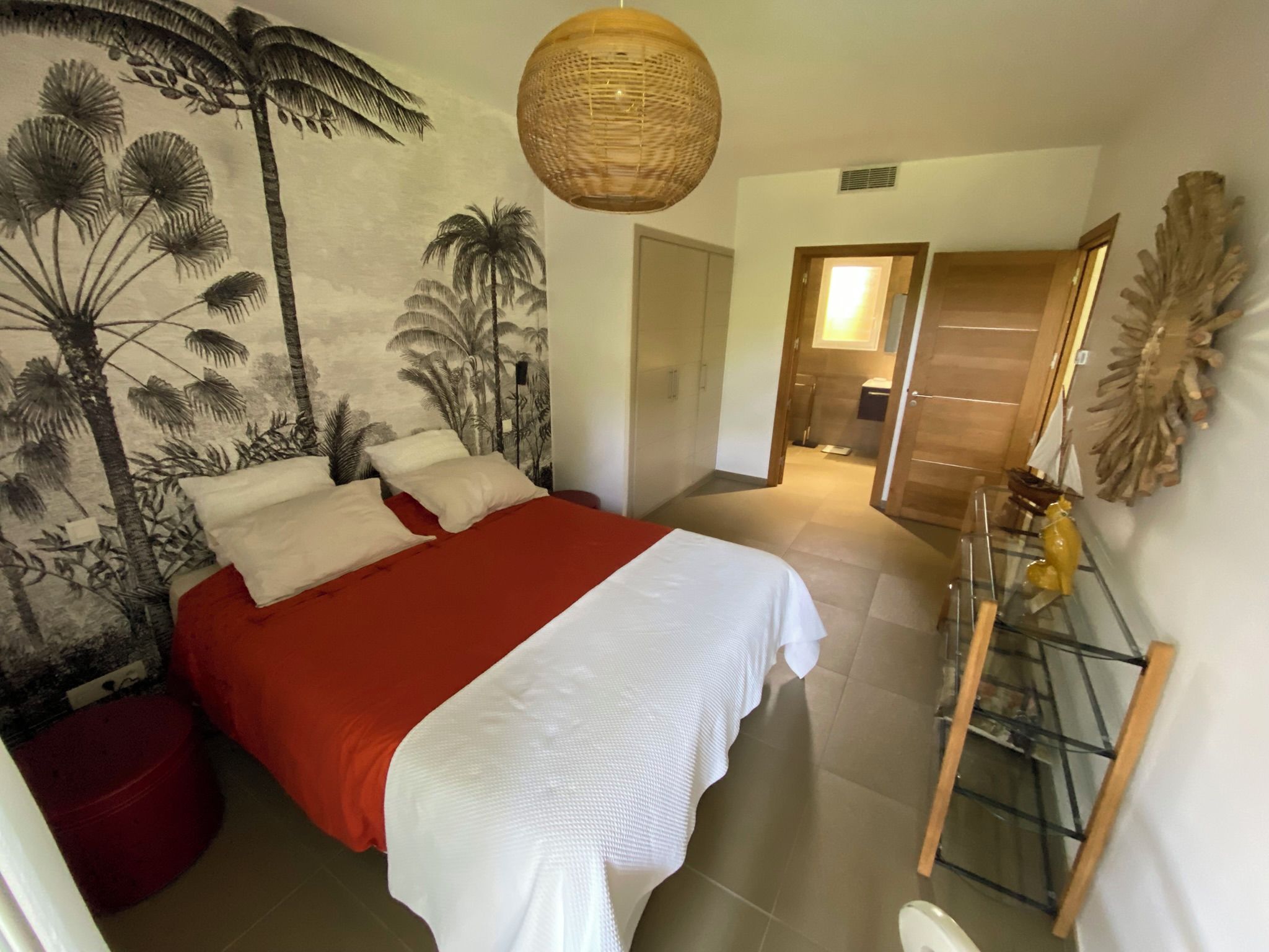 A bedroom with a bed, a wall mural, a hanging light fixture, and a bathroom visible through an open door.
