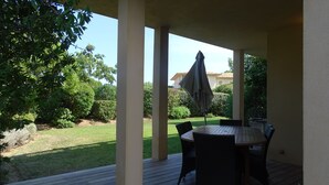 A covered patio with a table and chairs, surrounded by greenery and a view of a house.