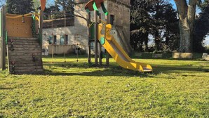 Un terrain de jeux avec un toboggan jaune et des structures d’escalade, situé devant un bâtiment en pierre et de grands arbres.
