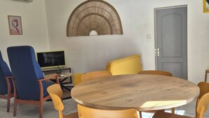 Un coin repas avec une table ronde en bois, des chaises jaunes, une chaise bleue, un téléviseur et un élément décoratif mural.