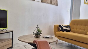 Un salon avec un canapé jaune, une table basse ronde en bois ornée d’une plante, un téléviseur posé sur un meuble noir et une œuvre d’art moderne accrochée au mur.