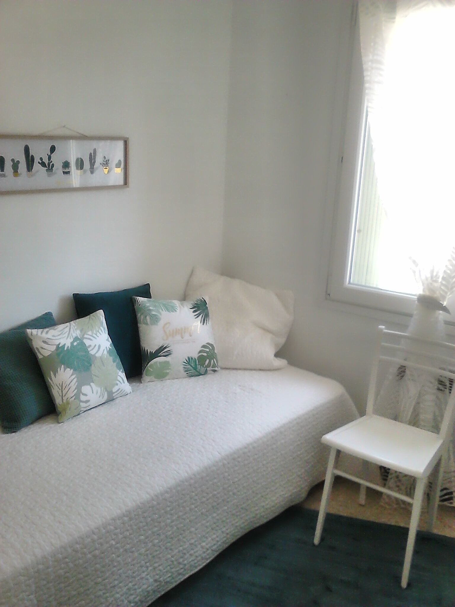 A bedroom with a bed, pillows, a small white chair, and a window with blinds.