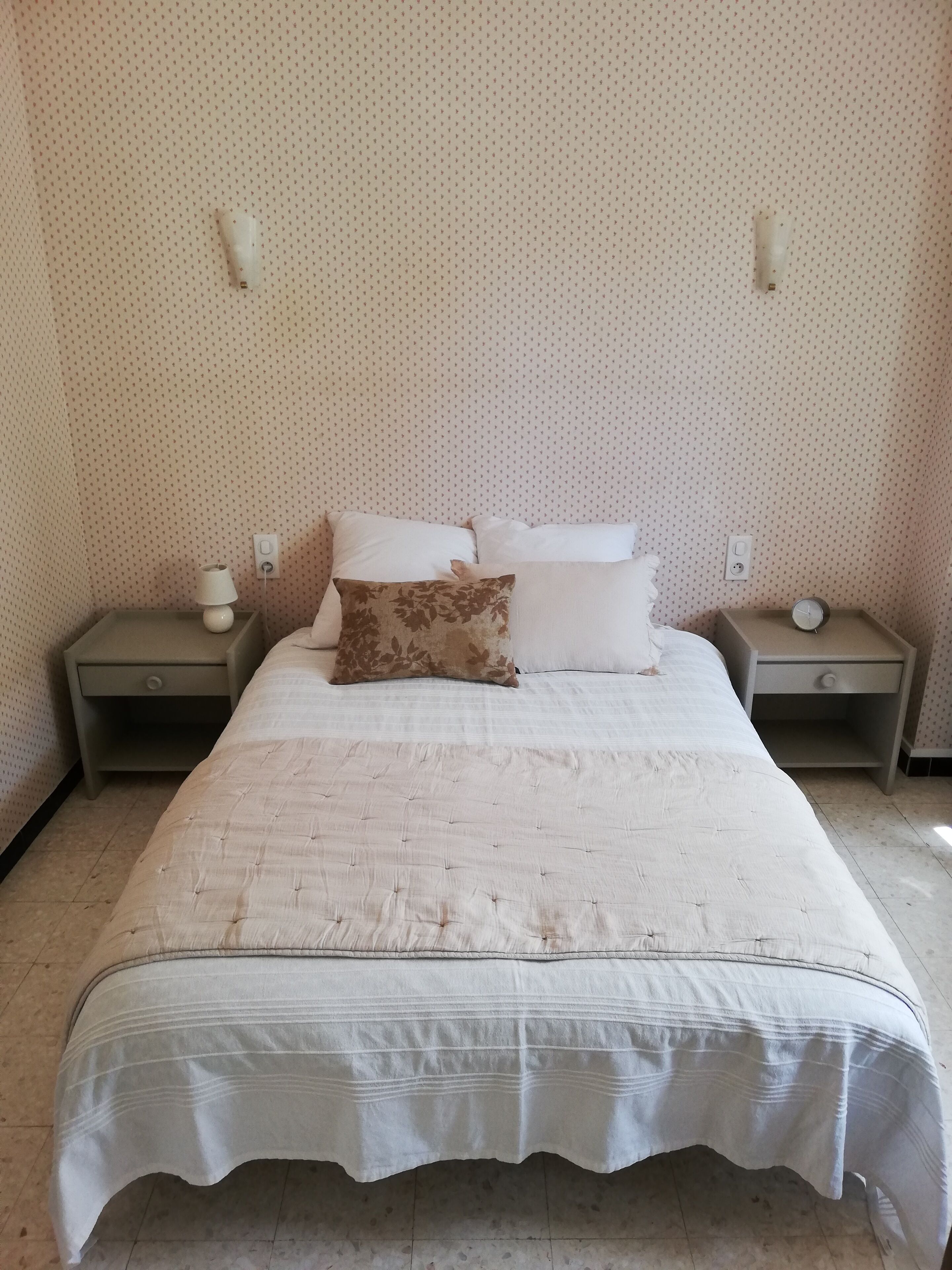 A neatly made bed with a white bedspread and two pillows, one with a leaf pattern. There are two bedside tables with lamps and a clock on one of them. The walls have a dotted pattern, and there is a power outlet on the wall.