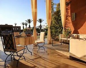 An outdoor patio area with a round table and chairs, a red building, and palm trees in the background. - Premium Suite for 2 in Marrakesh (Marrakech)