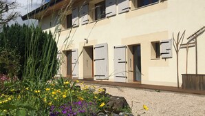 Un bâtiment comportant plusieurs pièces, chacune dotée d’une porte et de fenêtres. On aperçoit des plantes et des fleurs au premier plan.