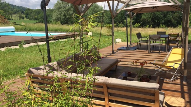 Un espace extérieur aménagé avec des meubles en bois, abrité par un auvent, une piscine en arrière-plan et une vue sur un paysage verdoyant.
