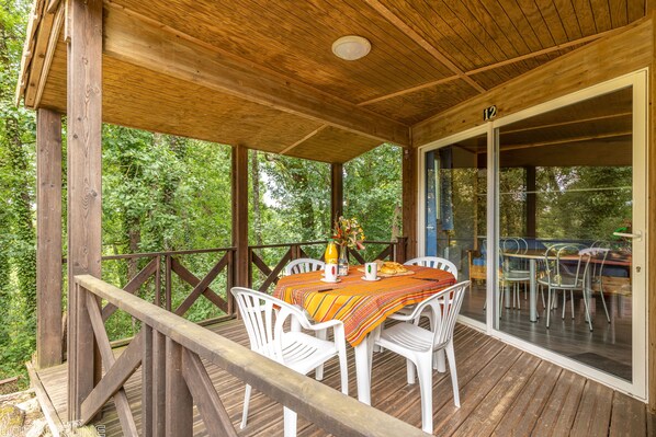 Ein überdachter Essbereich im Freien mit Holzdeck, weißen Kunststoffstühlen und einem Tisch, gedeckt mit einer Streifen-Tischdecke.