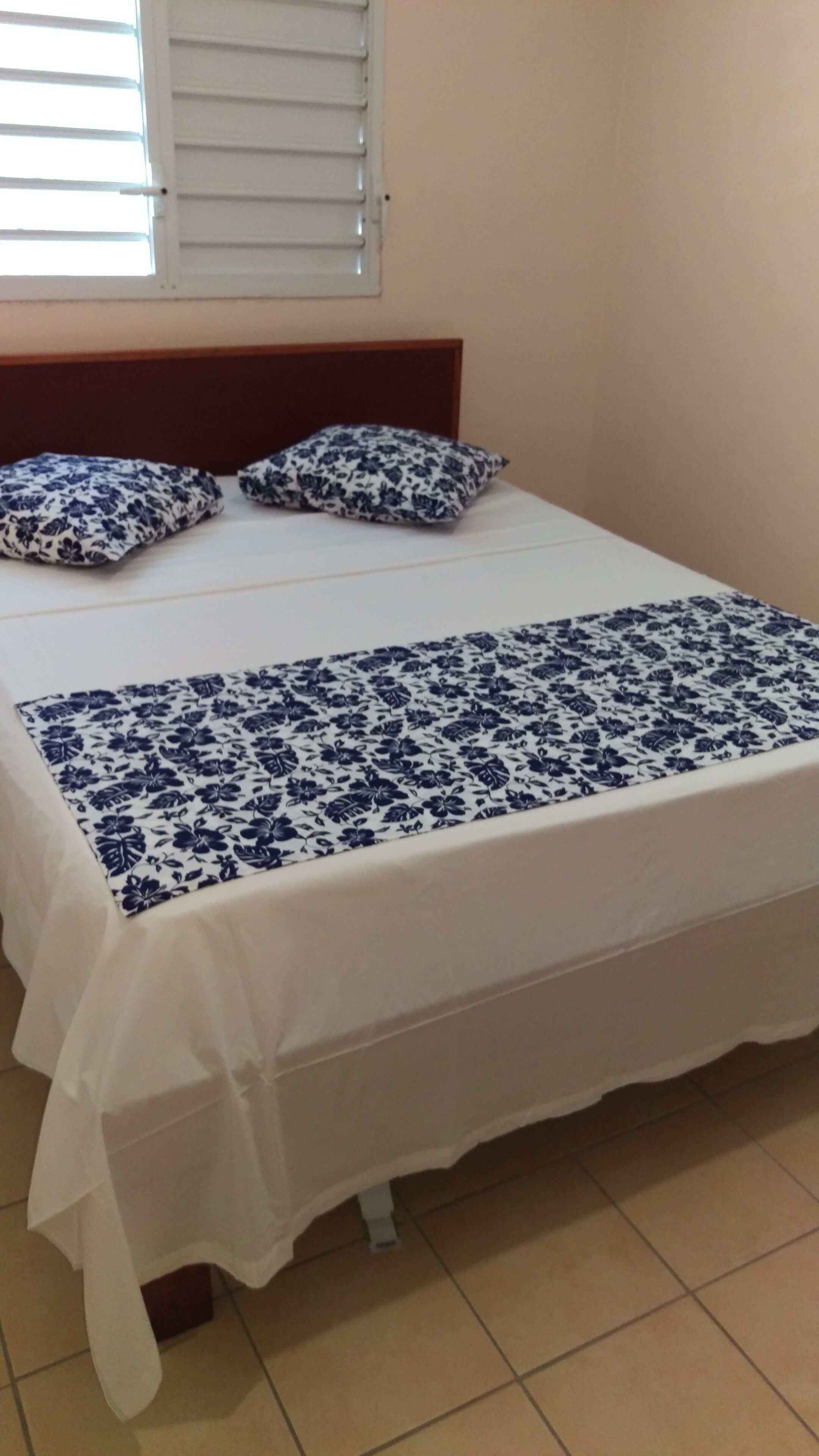 A bed with a white and blue floral bedspread, two matching pillows, and a wooden headboard in a room with beige walls and a window with blinds.
