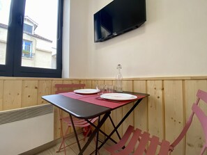 Une pièce avec une table dressée pour deux personnes, un téléviseur fixé au mur et une fenêtre donnant sur les bâtiments environnants.
