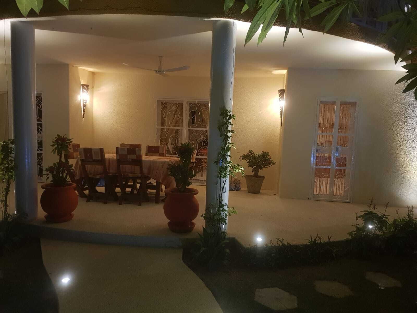 A covered outdoor dining area with a table and chairs, surrounded by potted plants and a ceiling fan.