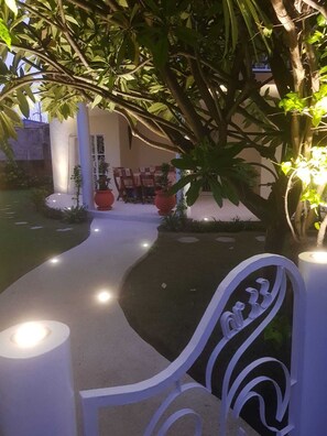 A garden area with a pathway leading to a seating area with chairs and tables, surrounded by potted plants and trees.